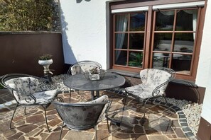 An outdoor patio with a round table and four chairs, a potted plant, and a window with curtains. - Holiday apartment with 1 bedroom (Walsdorf-Zilsdorf)