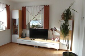 Ein Wohnzimmer mit einem Flachbildfernseher, einem Schrank, einem Topf mit einer Pflanze und einem Fenster mit Vorhängen.