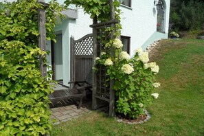 Uma pérgola de madeira coberta por plantas trepadoras e flores brancas, um banco de madeira e um edifício branco com um portão de treliça.
