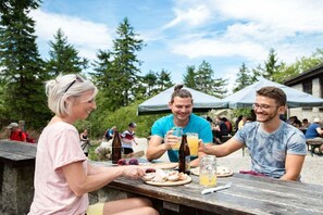 Eine Gruppe von Menschen sitzt an einem Tisch im Freien mit Essen und Getränken, umgeben von Bäumen und blauem Himmel.
