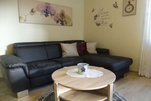 A living room with a dark leather sofa, a round wooden coffee table with a candle, and a wall with a floral painting and decorative butterflies. - Holiday apartment with 1 bedroom (Fichtelberg)
