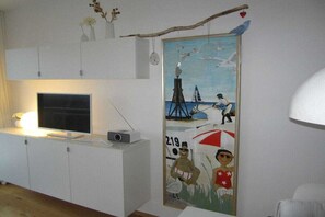 A room with a flat-screen TV on a white cabinet, a painting on the wall, and a white lamp. - Holiday apartment with 1 bedroom (Cuxhaven)