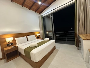Une chambre d’hôtel avec un grand lit, une table de chevet, un bureau et un balcon offrant une vue sur le ciel nocturne.