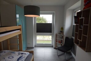 A room with a bunk bed, a desk with a chair, a window with blinds, and a bookshelf. - Holiday apartment with 2 bedrooms (Varel-Dangast)