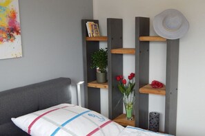 Ein Schlafzimmer mit einem Bett, einem grauen Sofa, einem an der Wand befestigten Regal mit Büchern, einer Topfpflanze, einer Vase mit Blumen und einem Hut.