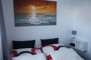 Une chambre à coucher avec un lit, des oreillers, une table de chevet, une lampe et un tableau représentant un coucher de soleil sur l’océan, accroché au mur.
