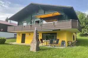 Ein zweistöckiges Gebäude mit Balkon, Außenbestuhlung und einer Steinsäule.