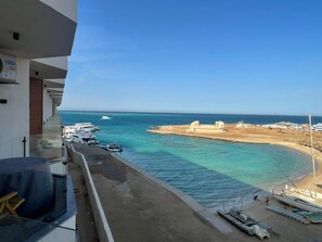 Ein Balkon mit Blick auf einen Strand und Boote, ein Sandbereich mit einem Gebäude, und ein klarer Himmel.