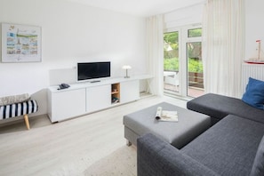 A modern living room with a grey sofa, a flat-screen TV on a white cabinet, a small table with a magazine, and a window with a view of greenery. - Holiday apartment in Juist with swimming pool (Juist)
