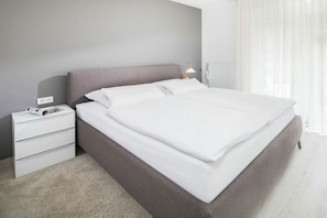 A neatly made bed with a grey headboard, a white bedside table with a lamp, and a white radiator in the background. - Holiday apartment in Juist with swimming pool (Juist)