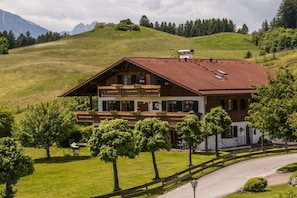 Ein zweistöckiges Chalet mit Balkon, umgeben von einem gepflegten Rasen und Bäumen, vor einer Bergkulisse.