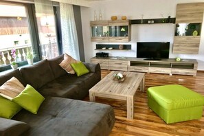 A modern living room with a sofa, coffee table, and ottoman, wooden flooring, and a TV on a wooden stand. - Holiday home with 5 bedrooms (Steinwiesen)