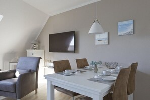A modern dining area with a white table, brown chairs, a flat-screen TV mounted on the wall, and a painting above the table. - Holiday apartment with 1 bedroom (Cuxhaven)