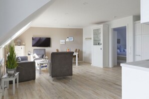 Een moderne woonkamer met een flatscreen televisie, grijze banken, een salontafel, en een eethoek met een wit kastje en een glazen vitrine.