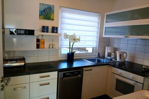 A modern kitchen with white cabinets, a dishwasher, a coffee maker, and a window with blinds. - Holiday home Lechseeferien II (Lechbruck am See)