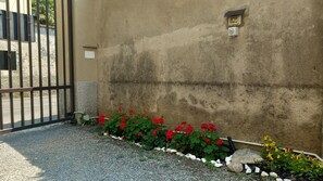 Une entrée fermée par un portail et un mur en béton, un chemin de gravier et un parterre de fleurs rouges.