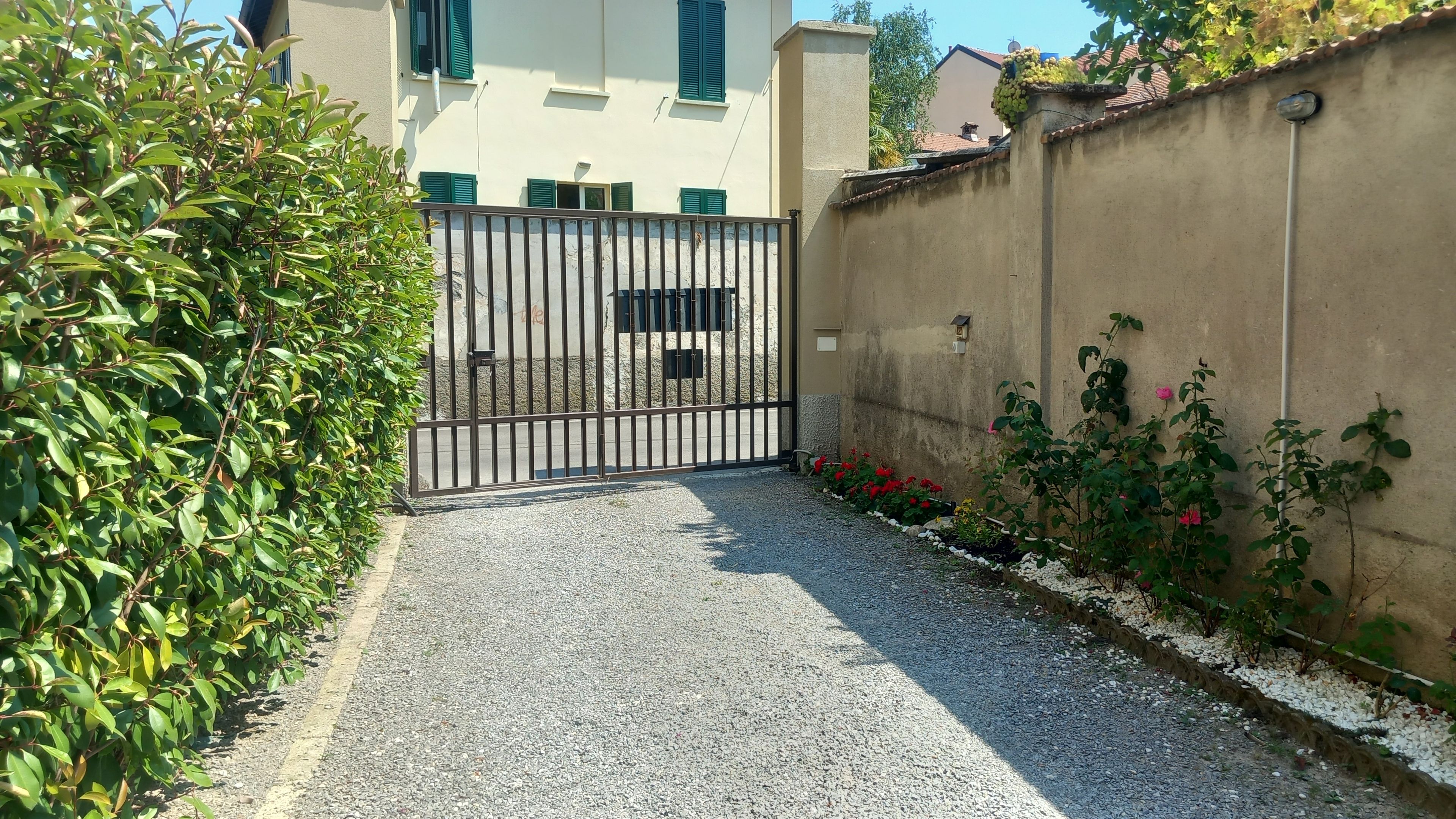 Une entrée fermée par un portail donne sur un chemin de gravier qui mène à un bâtiment à la façade beige et aux volets verts.