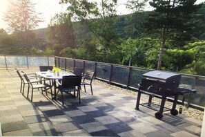 Um pátio externo com mesa e cadeiras, churrasqueira e vista para árvores e colinas.