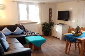 Ein Wohnzimmer mit einer Couch, einem Fernseher, einem Esstisch mit Früchten und einer Zimmerpflanze.