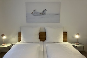 Un lit double avec une literie blanche, deux lampes de chevet et un tableau représentant deux lapins, accroché au mur.