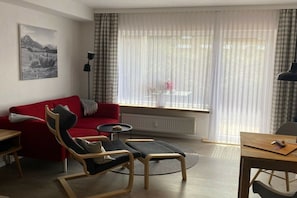 A modern living room with a red sofa, a wooden chair, a small round table, and a view through sheer curtains. - Alpenflair holiday apartment 235 (Oberstdorf)