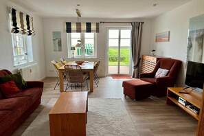 A living room with a sofa, a wooden coffee table, a TV stand, and a dining area with chairs and a table. - Holiday home in Norddorf with swimming pool (Norddorf)