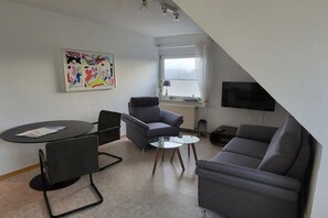 A modern living room with a sofa, armchair, and coffee table. There is a painting on the wall and a TV mounted on the wall. - Holiday apartment with 1 bedroom (Neuharlingersiel)