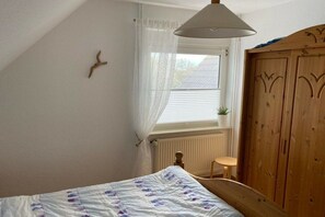 A bedroom with a bed, a window with curtains, a radiator, a wooden wardrobe, and a small stool. - Holiday apartment with 1 bedroom (Neuharlingersiel)