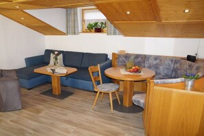 A cozy room with a blue sofa, a wooden dining table with chairs, and a small window with curtains. - Holiday apartment with 2 bedrooms (Obermaiselstein)