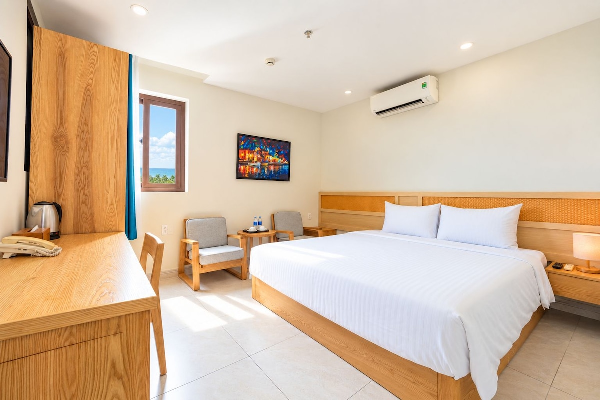 A hotel room with a bed, a desk, two chairs, a nightstand, an air conditioner, and a painting on the wall.