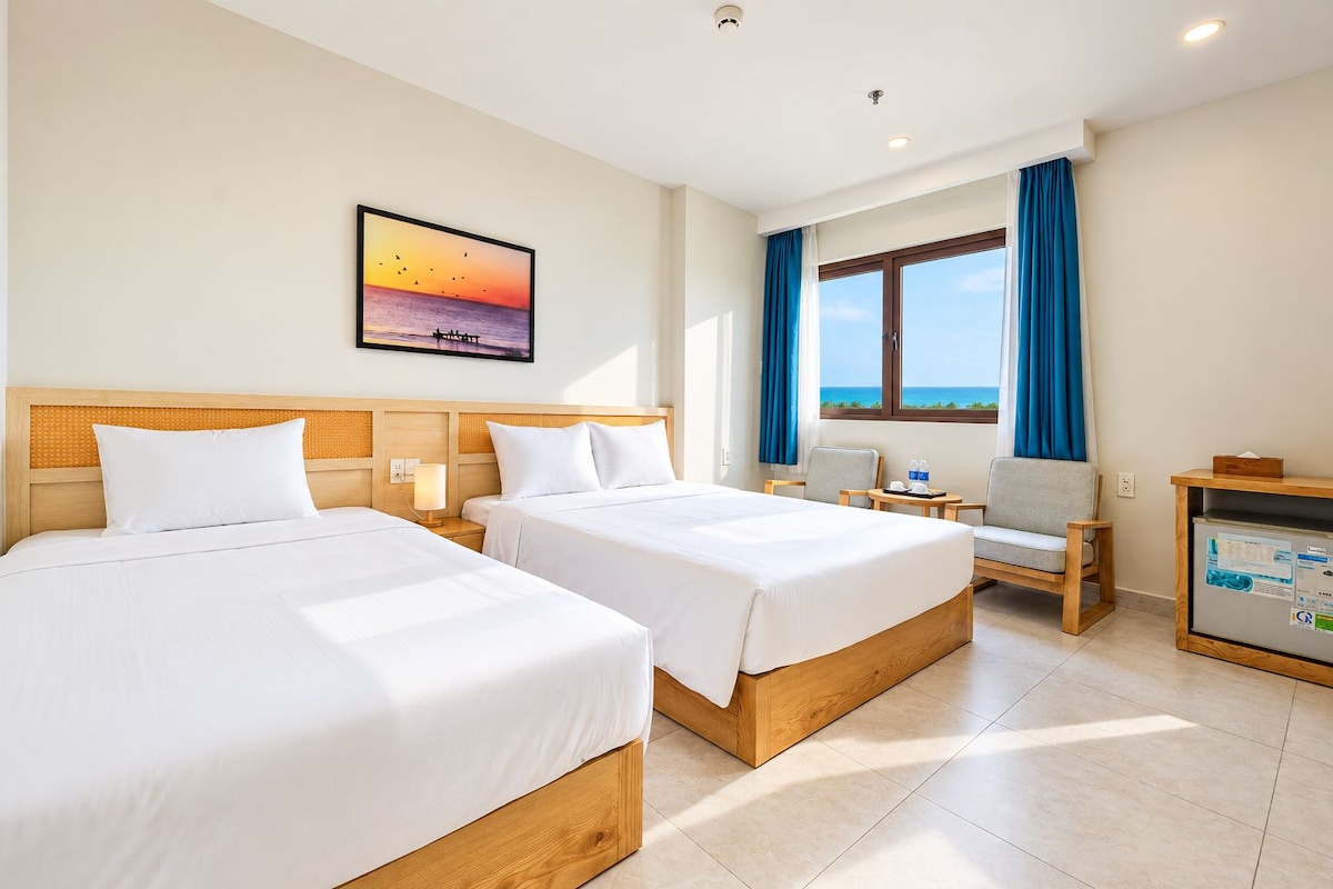 A hotel room with two beds, a desk, a chair, a window with a view, and a wall-mounted picture.