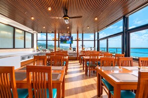 Un restaurante con mesas y sillas de madera, una pantalla de televisión y amplios ventanales con vistas al océano.
