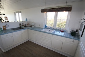 A kitchen with white cabinets, a blue-tiled countertop, a sink, and a window with a view of greenery outside. - Top Wadden Sea holiday home (Esbjerg)