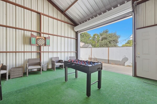 A room with a foosball table, two wicker chairs, a dartboard, and a green artificial turf floor.