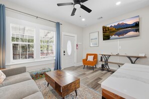 A modern living room with a sofa, a wooden coffee table, a small orange armchair, a TV mounted on the wall, and a painting of a mountain landscape.