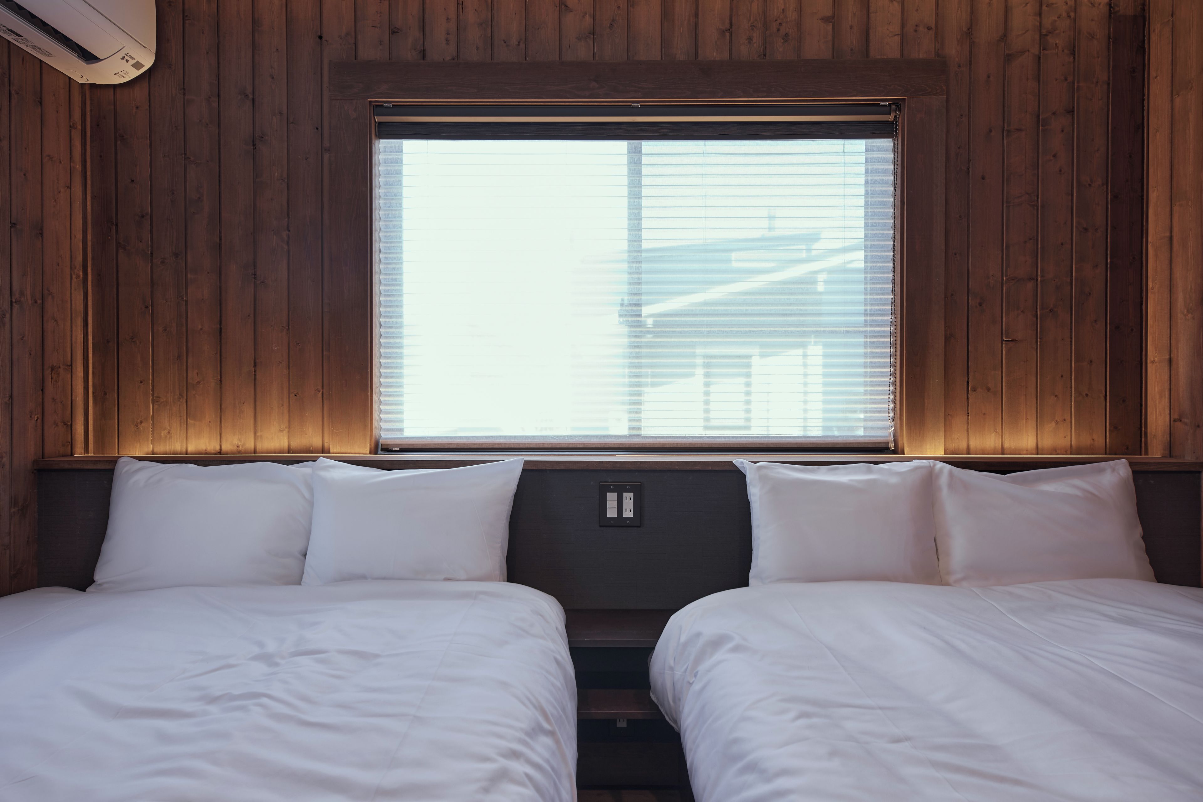 A room with two beds, wooden walls, a window with blinds, and a thermostat on the wall.