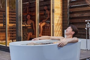 A person relaxing in a large, round hot tub with a view of a wooden building and a glass wall reflecting another person.