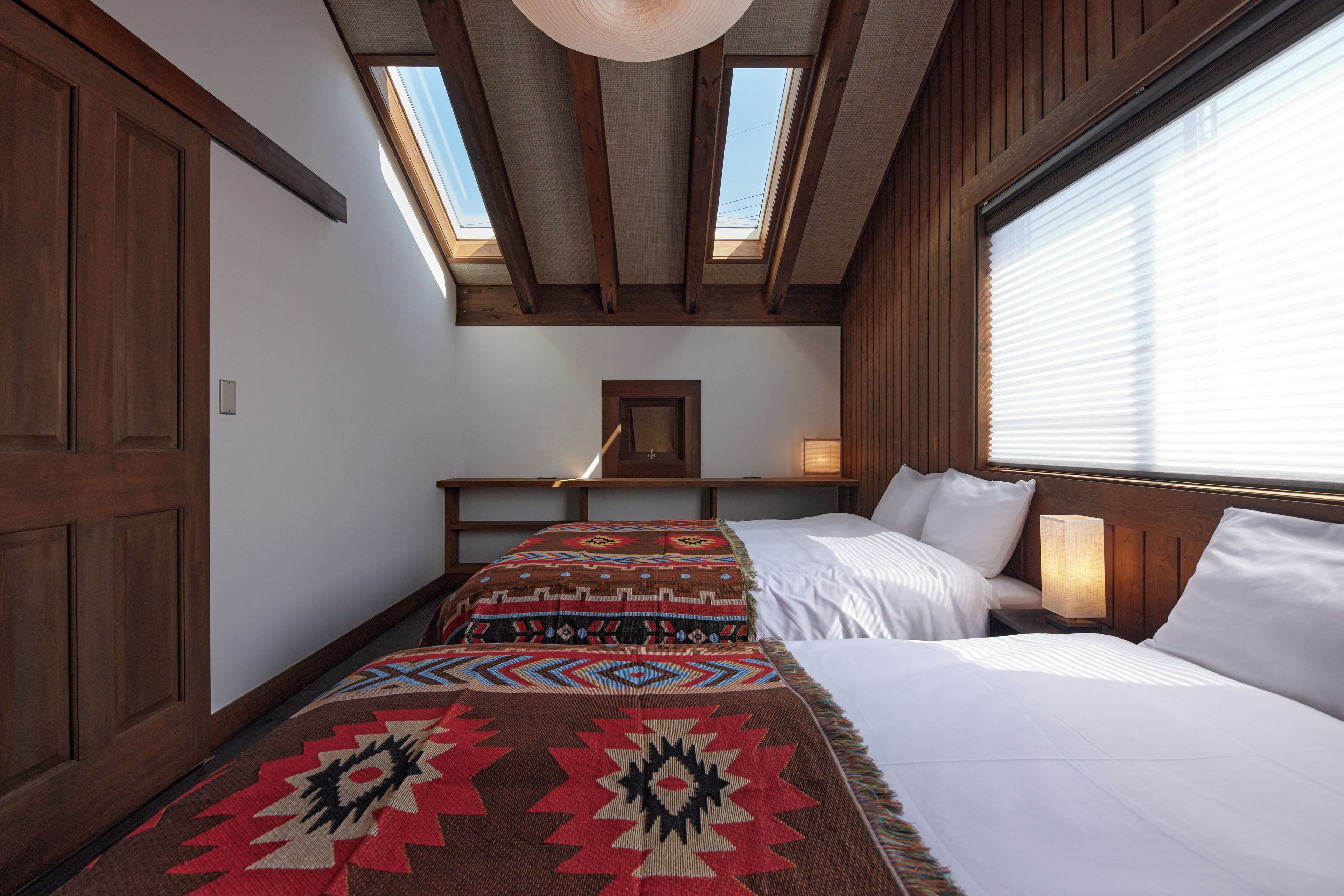 Un dormitorio con tragaluces, una cama con colcha estampada, una mesita de noche con lámpara y una ventana con persianas.