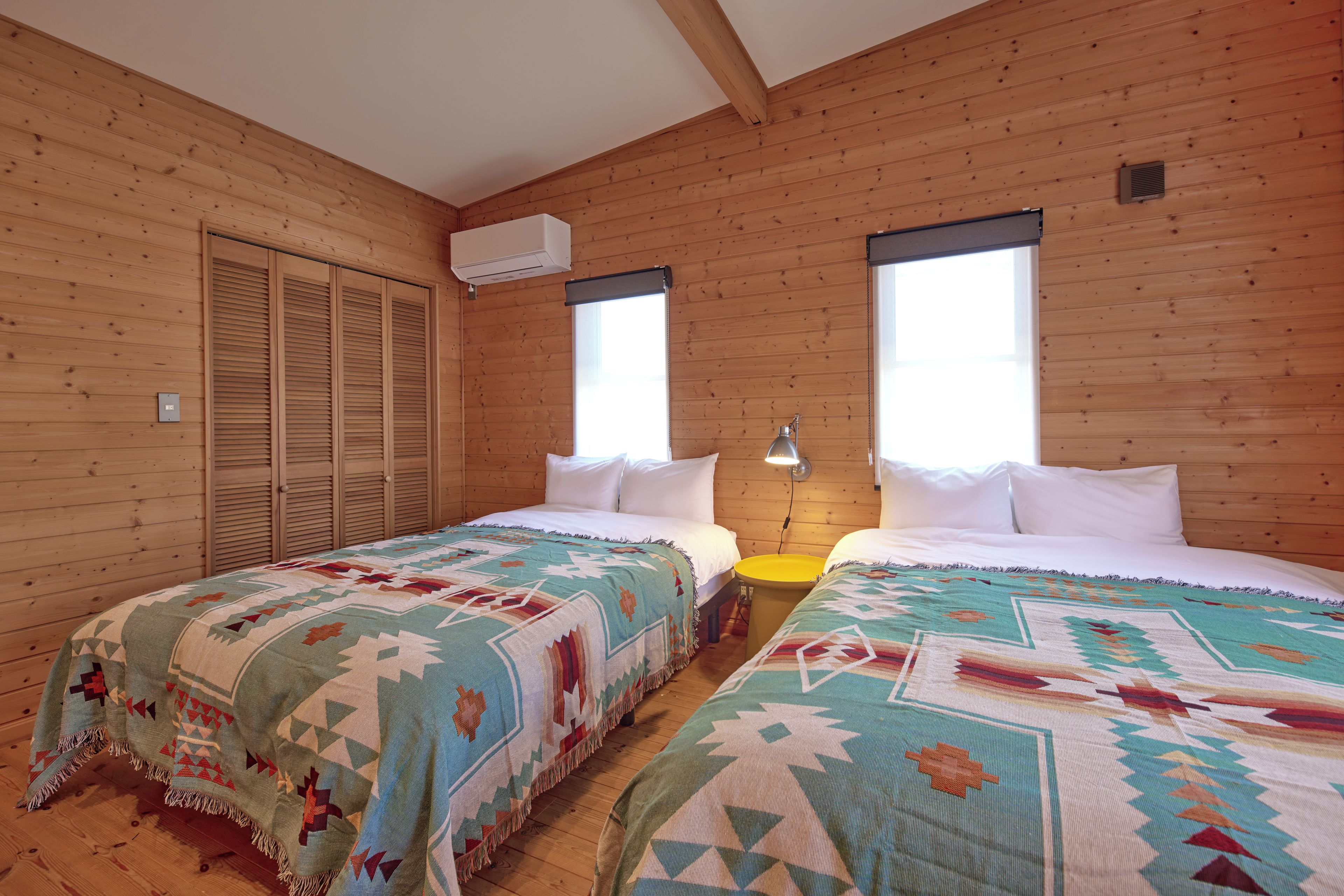 A room with two beds, wooden walls, a window, and an air conditioning unit.