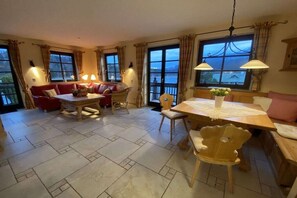 Ein gemütliches Wohnzimmer mit rotem Sofa, Esstisch aus Holz und Stühlen, großen Fenstern mit Vorhängen und Blick aufs Meer.