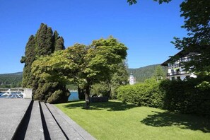 Ein gepflegter Garten mit einem Weg, der zu einem Gebäude mit einem Balkon führt, der auf ein Gewässer und Hügel im Hintergrund blickt.