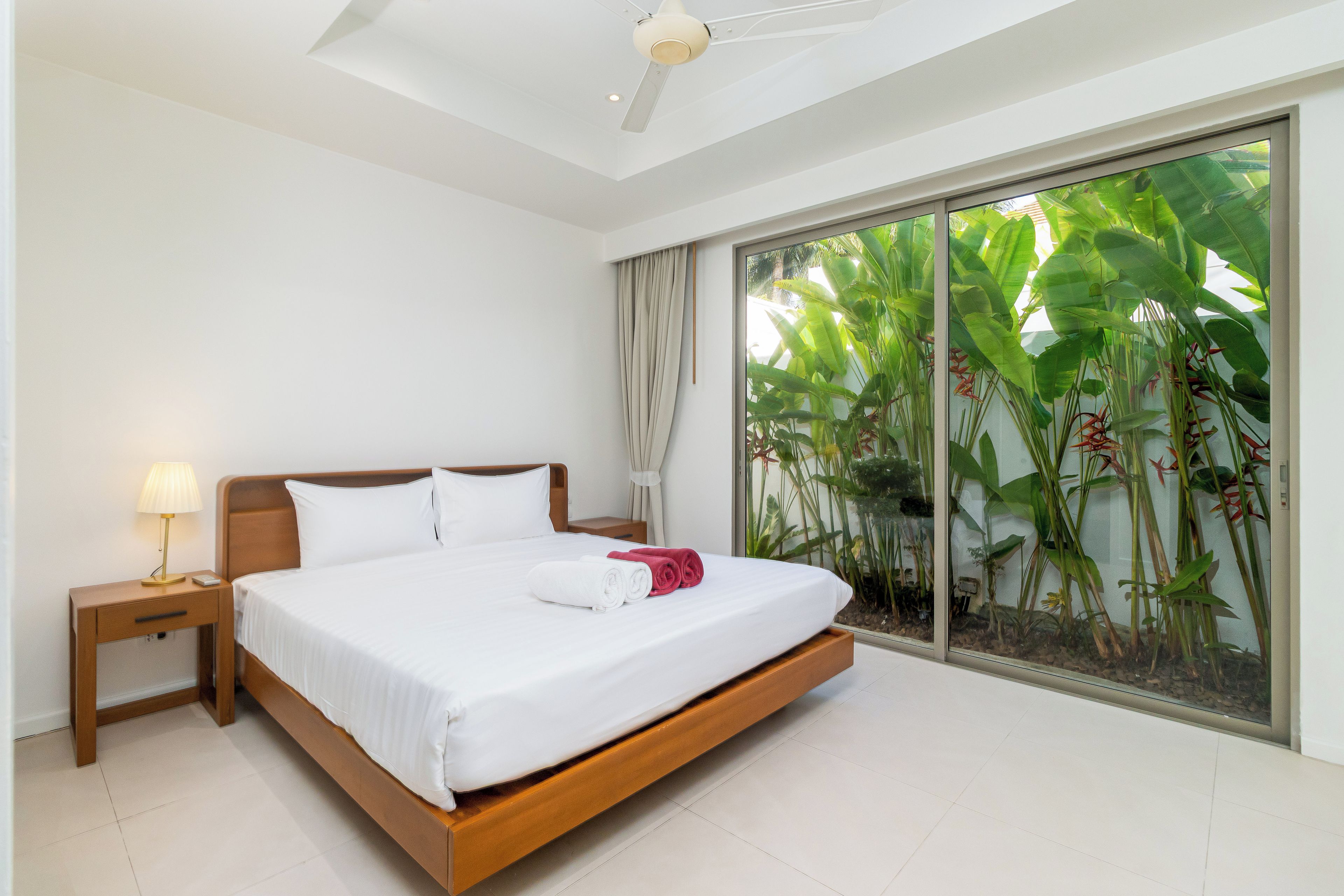 Habitación de hotel con una cama grande, una mesita de noche, una lámpara y vistas a plantas tropicales a través de un ventanal corredero.