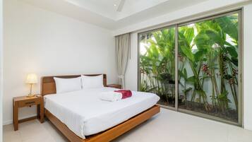 Habitación de hotel con una cama grande, una mesita de noche, una lámpara y vistas a plantas tropicales a través de un ventanal corredero.