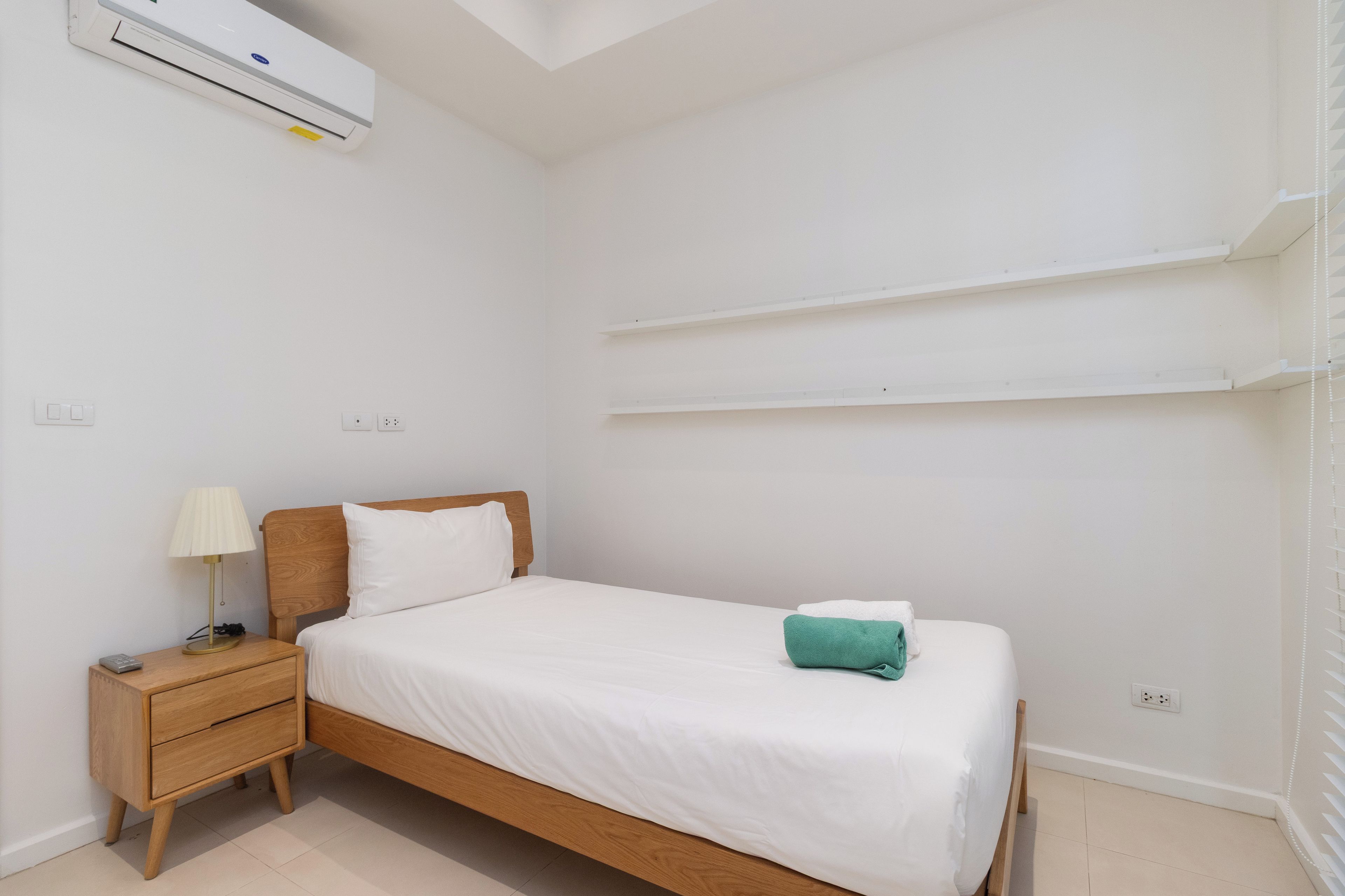 Habitación individual con cama de una sola plaza, cabecera de madera, mesita de noche con lámpara y aire acondicionado instalado en la pared.