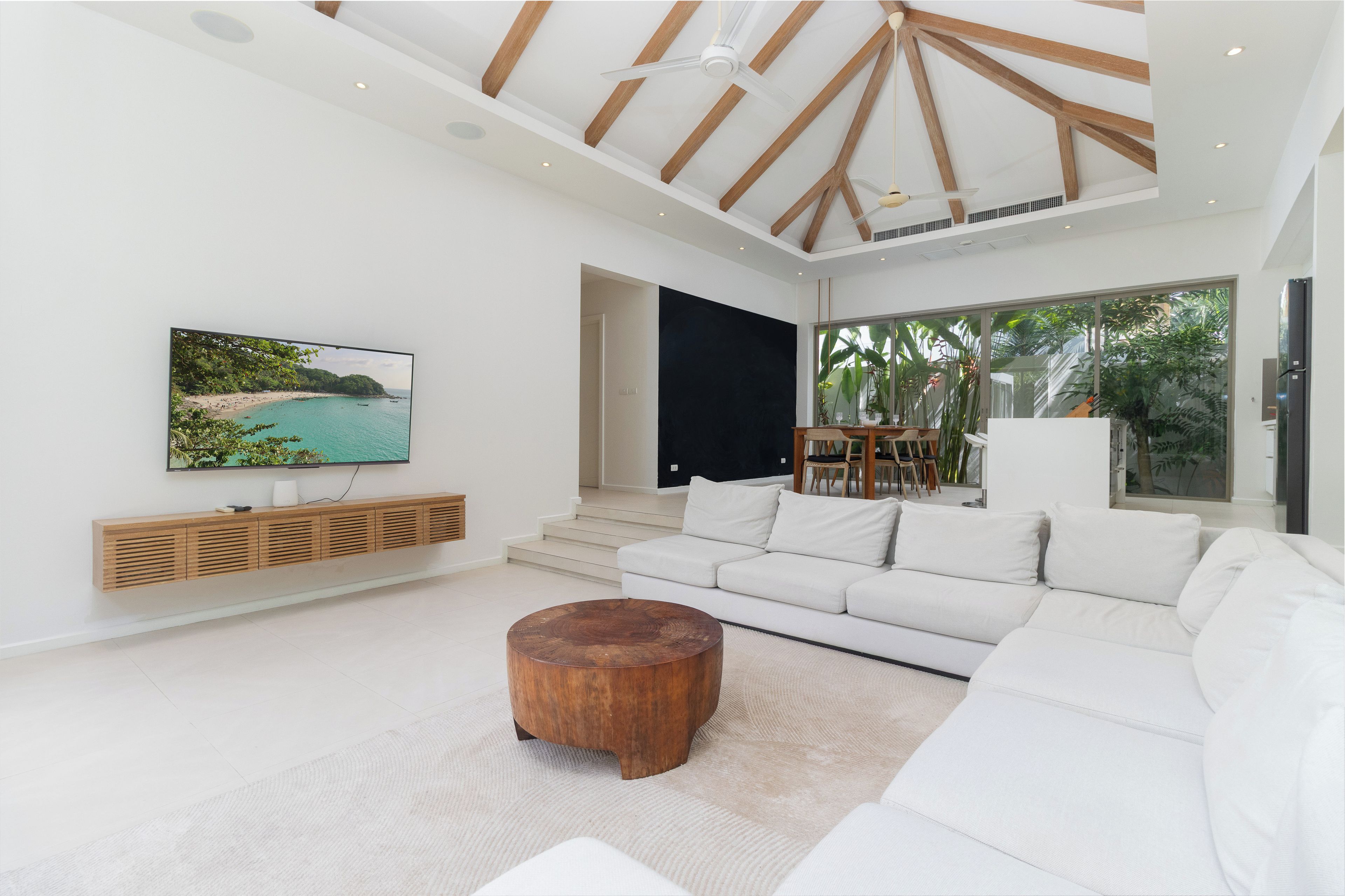 Una sala moderna con un televisor de pantalla plana grande, sofás blancos, una mesa de centro de madera y vistas a una playa tropical a través de amplias ventanas.