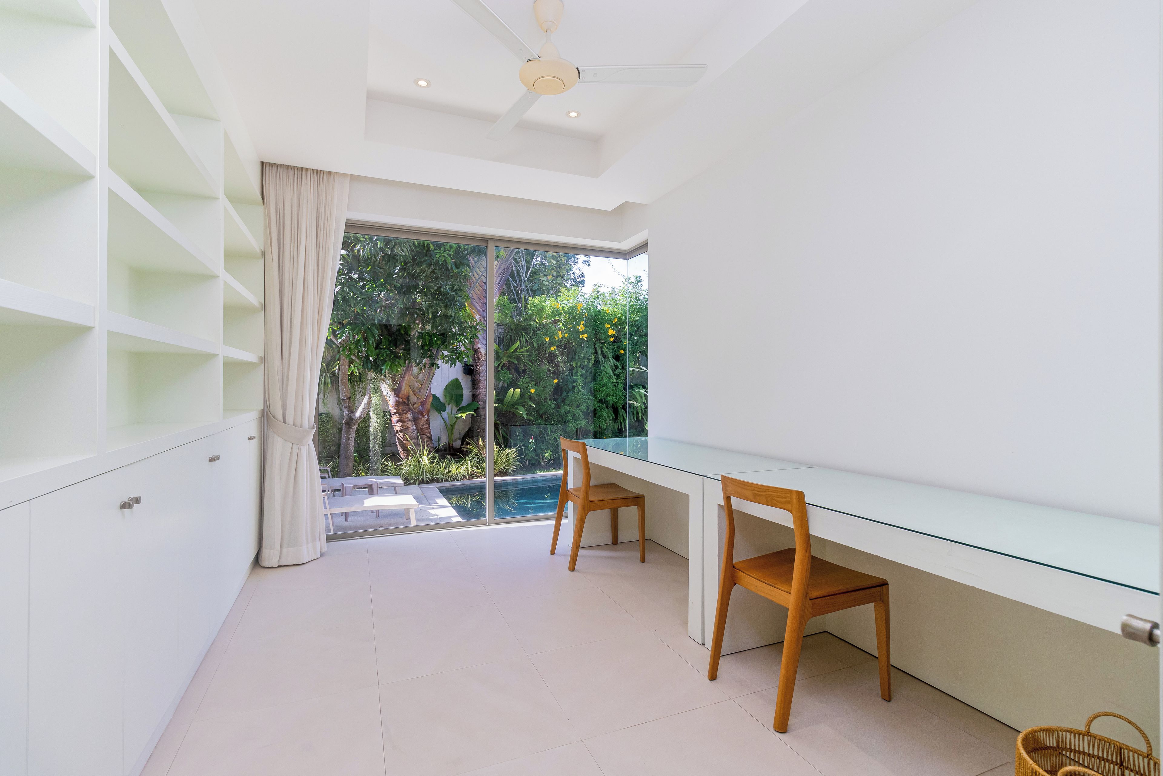 Una habitación moderna con un escritorio de cristal, dos sillas de madera, un ventilador de techo y vistas a un jardín a través de un ventanal grande.