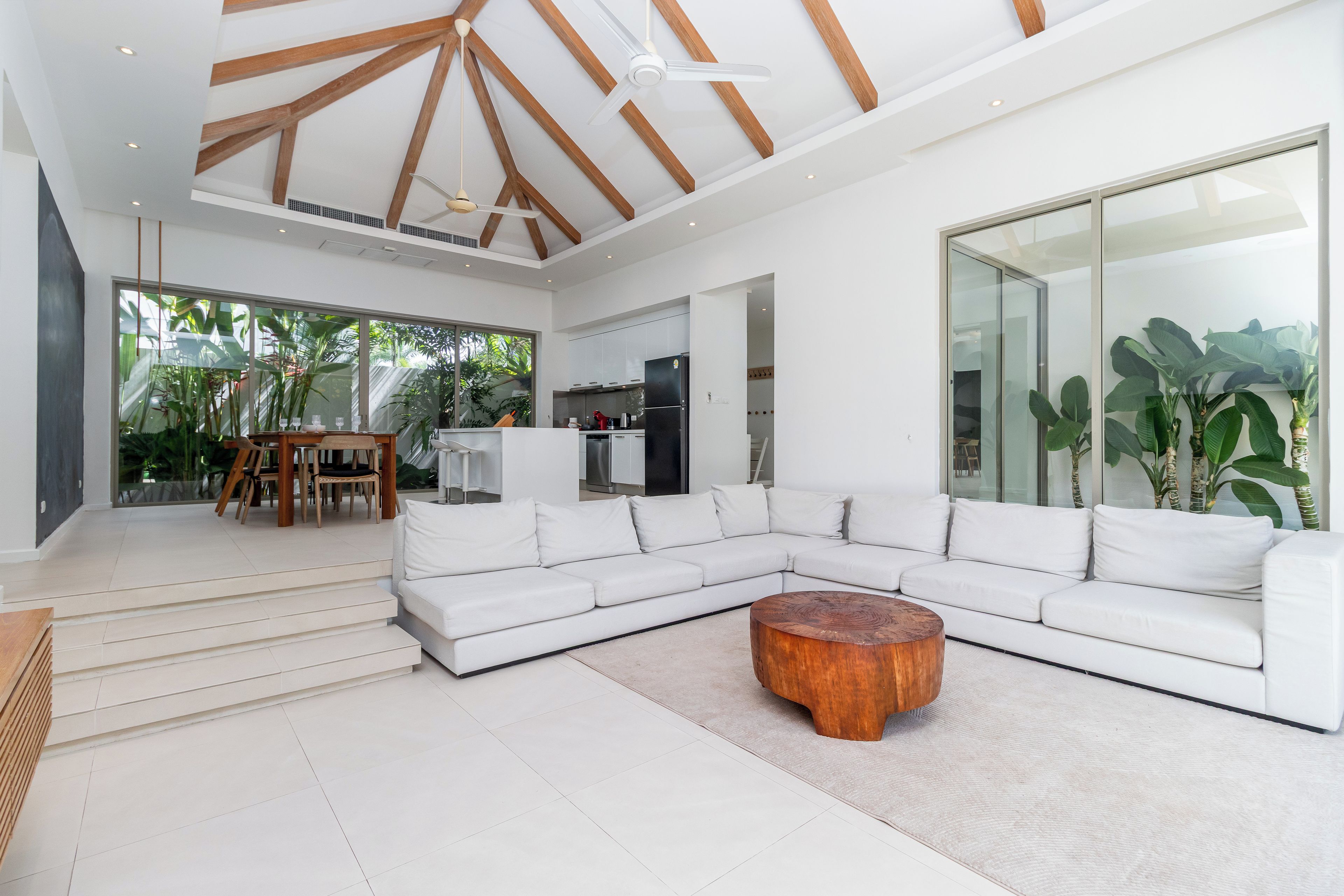 A spacious living room with a white sectional sofa, a wooden coffee table, and a large window offering a view of the outdoors.