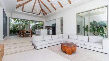 A spacious living room with a white sectional sofa, a wooden coffee table, and a large window offering a view of the outdoors.