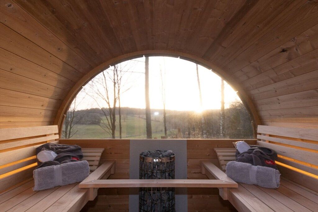 A wooden sauna with benches and a central fire pit, a large arched window, and a view of the outdoors.