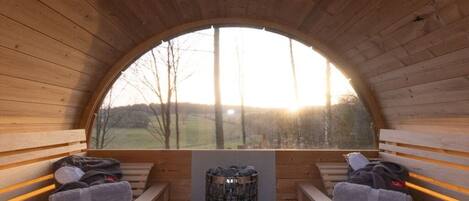 A wooden sauna with benches and a central fire pit, a large arched window, and a view of the outdoors.